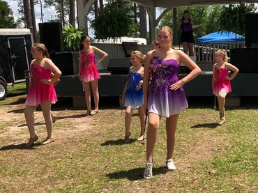 Young girls in colorful dance costumes pose in front of a gazebo outdoors.