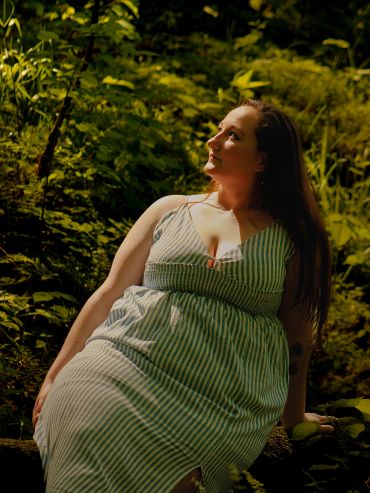 Woman in a striped dress sitting in a lush green forest, looking contemplative.