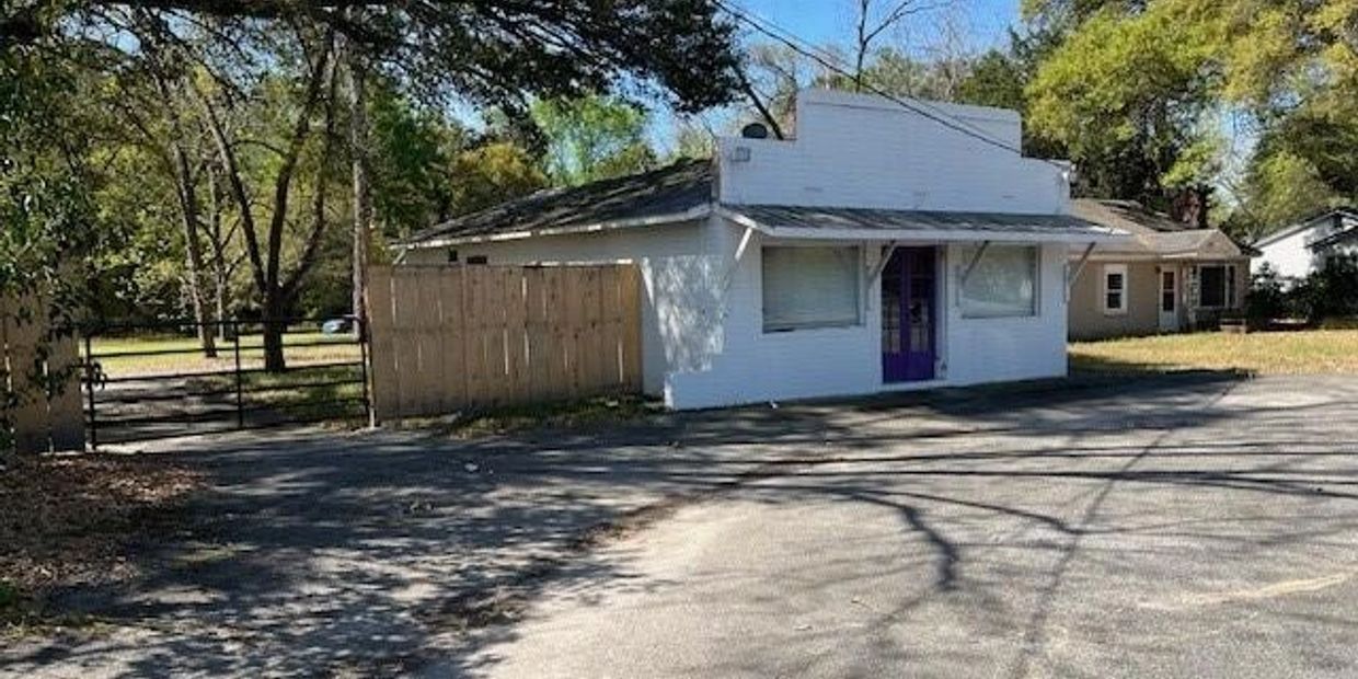 Small white building with purple door surrounded by trees and a paved area.