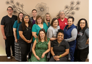 A diverse group of twelve people posing indoors against a beige wall with metal flower decorations.