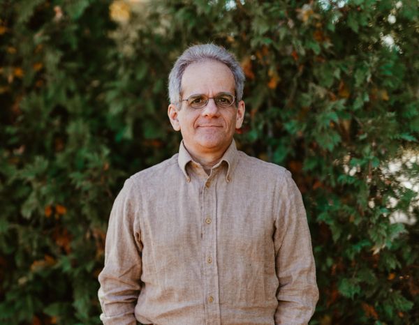 Man standing outdoors with greenery in the background, wearing glasses and a beige shirt.