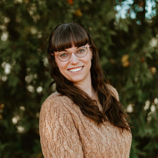 Smiling woman with glasses and long hair in a cozy brown sweater outdoors.
