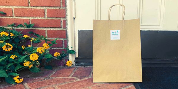 A brown delivery bag with a Priscilla's label on a doormat beside a floral bush.