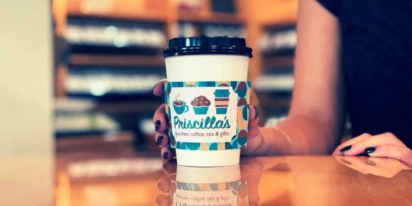 A hand holding a coffee cup with Priscilla's branded cup sleeve on a reflective table.