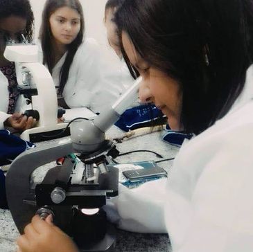 Student using a microscope in a science lab with peers in the background.