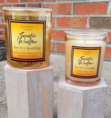 Two Sweater Weather scented candles on wooden stands against a brick wall.