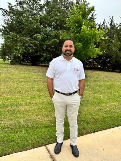 A man in a white polo and beige pants stands on grass near trees.