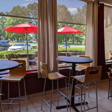 Cozy cafe seating with tall tables and wooden chairs by large windows overlooking outdoor umbrellas.