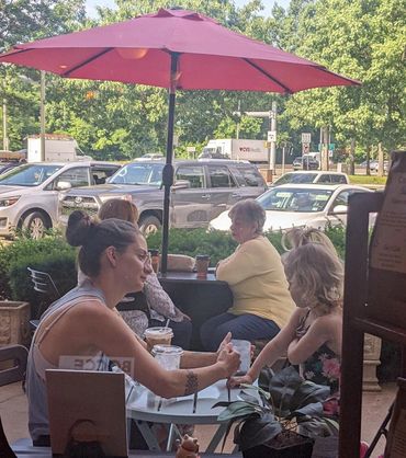 Two women sitting under a red umbrella at a cafe table, one showing something on her phone.
