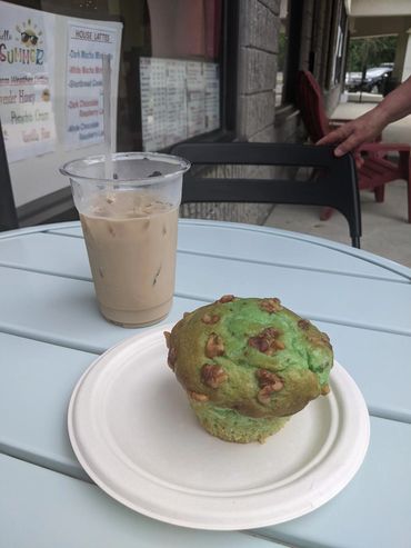 Iced coffee and a green muffin with walnuts on a white plate outside a cafe.