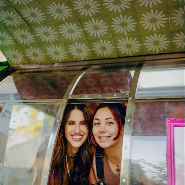 Two smiling women leaning out of a window with floral ceiling pattern above.