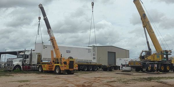 Two cranes lifting large containers near trucks and a warehouse under cloudy skies.