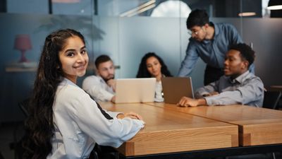 Young woman smiling in a meeting with colleagues working on laptops.