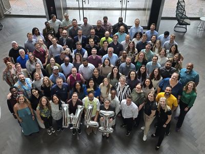 Large group photo of diverse people holding silver balloons forming 'WLT'.