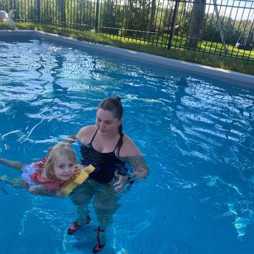 A woman helps a young girl swim in a clear outdoor pool on a sunny day.