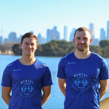 Two men in blue shirts standing outdoors near water with city skyline behind.