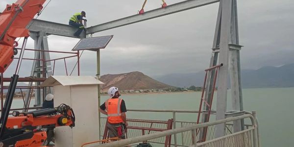 Workers assembling a large metal structure by the water using a crane.