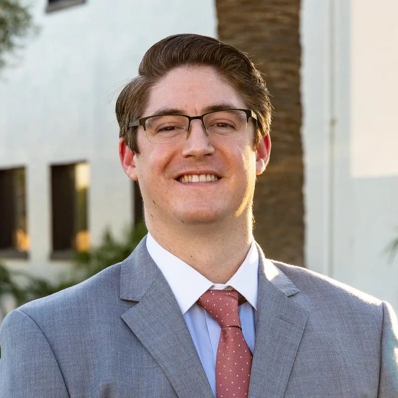 A smiling man in a gray suit and red tie stands outdoors.