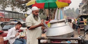 Street vendor serving chai near a colorful umbrella in a busy market area.