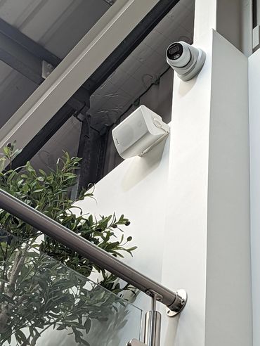 Security camera and speaker mounted on a white wall near a plant and railing.