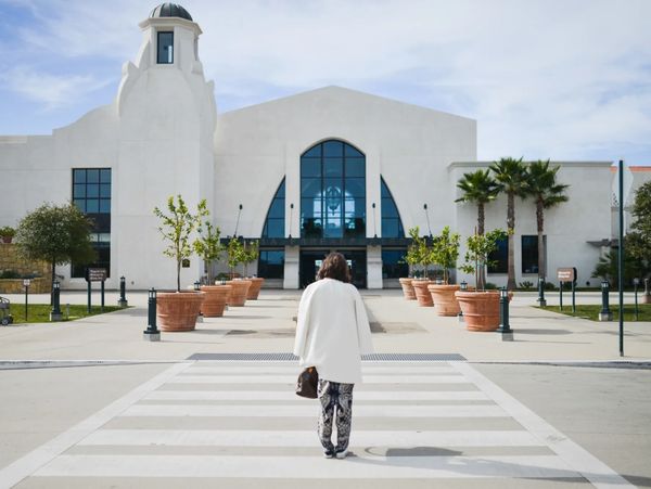 Person crossing a street towards a white building with potted plants.