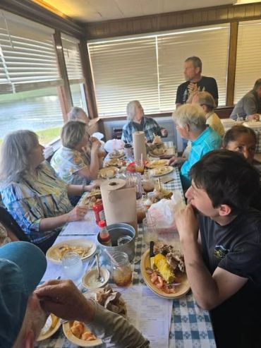 Group of people enjoying a meal together at a long, checkered table by a window.