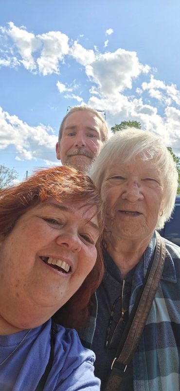 Three people smiling outdoors under a bright blue sky with clouds.