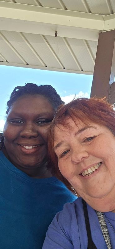 Two women smiling together under a covered outdoor area on a sunny day.