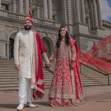A couple in traditional Indian wedding attire stands before an ornate building.
