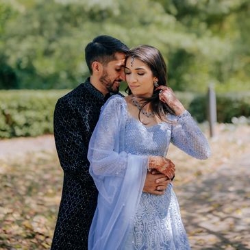 A couple embraces outdoors in traditional attire, surrounded by greenery.