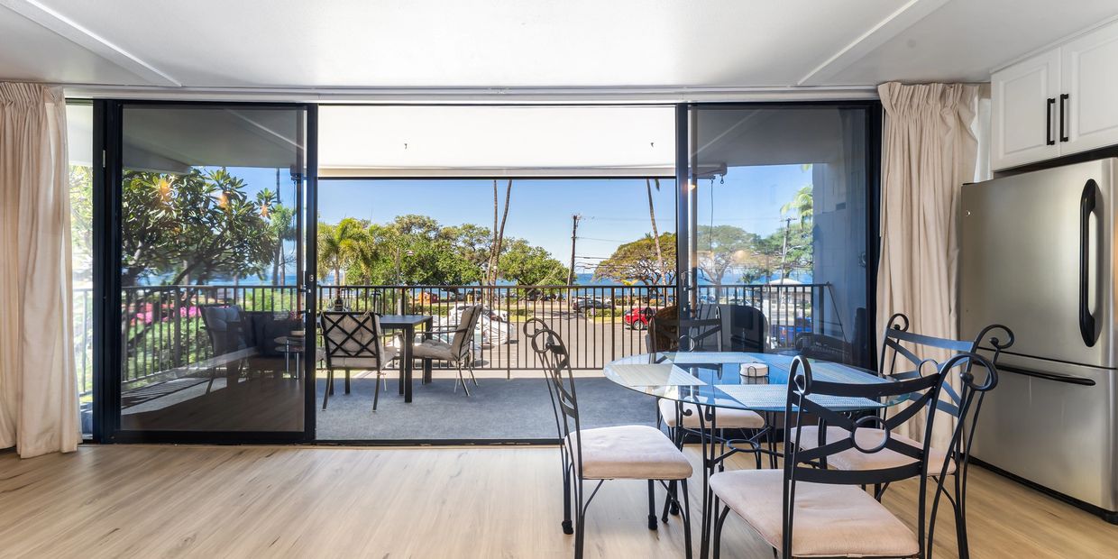 Bright dining area with glass table and balcony overlooking the ocean and palm trees.