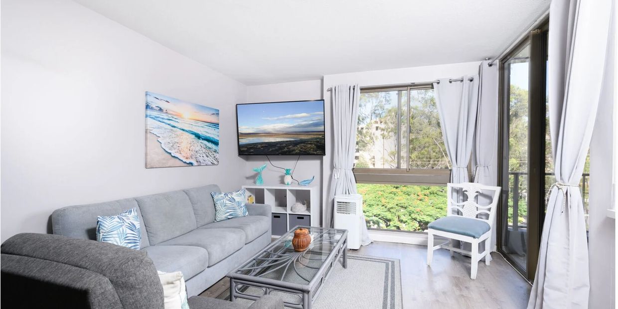 Bright living room with gray sofa, armchair, glass coffee table, and large windows with white curtains.