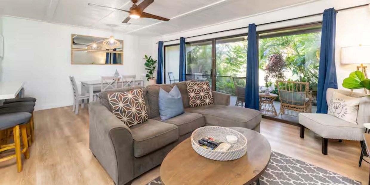 Bright living room with gray sofa, wooden coffee table, and large windows opening to a lush garden patio.