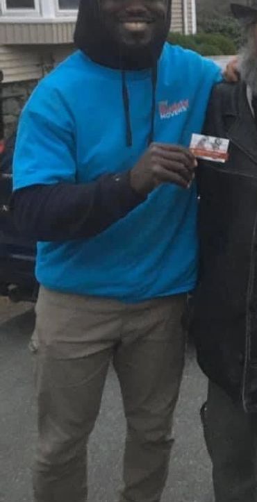 Two men smiling together outdoors, one wearing a blue hoodie and the other a black leather jacket and hat.
