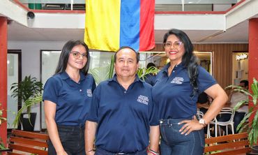 Three people in matching navy blue shirts pose indoors under a colorful flag.
