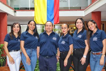 Group of six smiling people in navy blue shirts standing indoors under a Colombian flag.