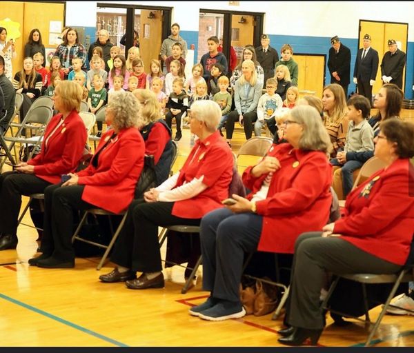 Elderly women in red jackets seated, children and adults watching in a school gym.