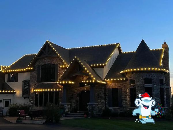A large house decorated with warm white string lights along the roof edges at dusk.