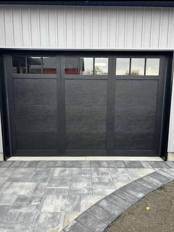Modern black garage door with upper windows and gray stone driveway.