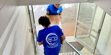Two movers in blue shirts carrying a wooden table up a carpeted staircase.