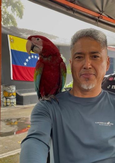 Hombre con guacamayo rojo en el hombro con bandera de Venezuela al fondo.