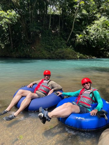 Two people relaxing on blue inner tubes in a clear river, wearing red helmets and life jackets.