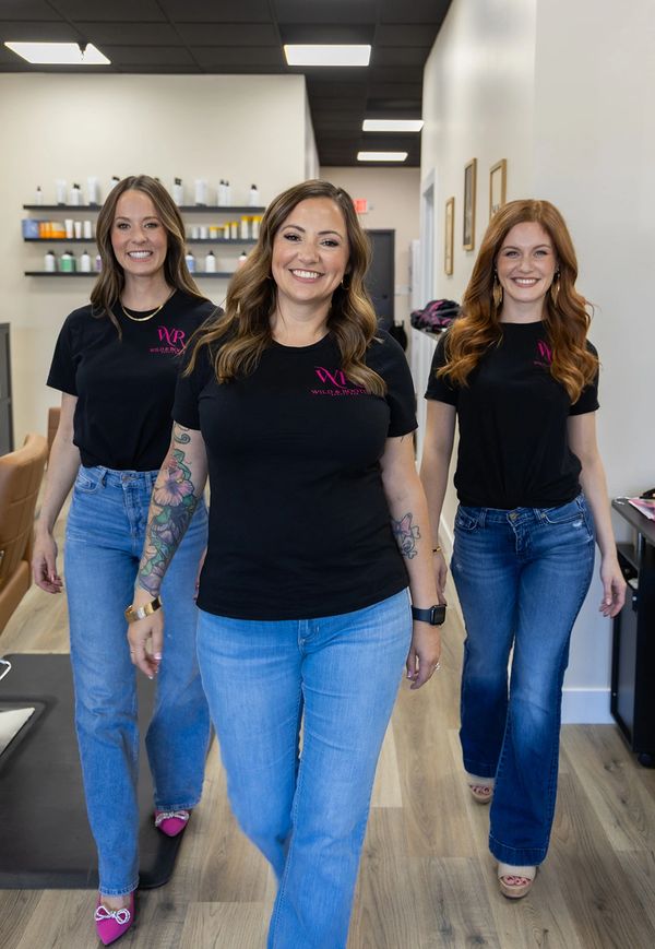 Three smiling women in black shirts and jeans walking confidently indoors.