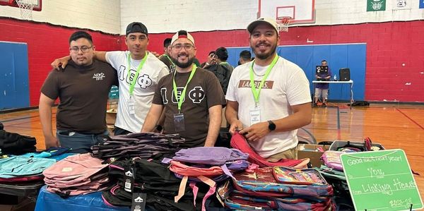Four men smiling behind a table of backpacks in a gym.