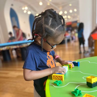 A child with braided hair plays with colorful building blocks at a green table indoors.