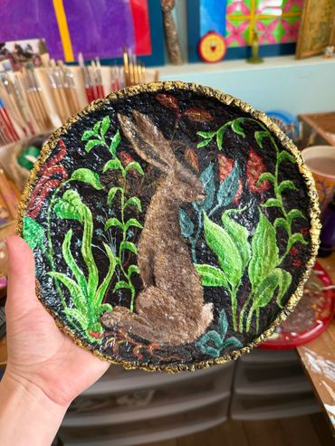 Papier mache plate painted with a rabbit and vibrant garden plants over a black background.