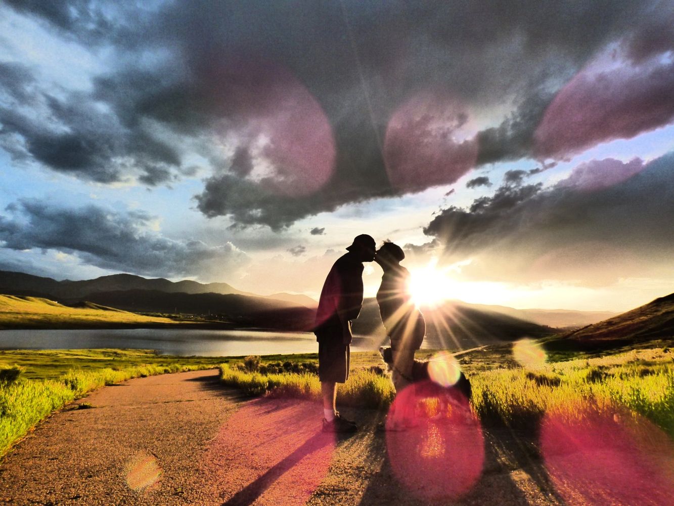 Silhouetted couple kissing at sunset on a scenic path by a lake and mountains.