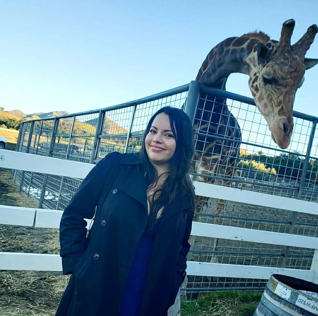 A woman smiling in front of a giraffe at an outdoor enclosure.