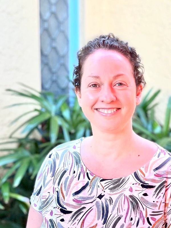 Smiling woman in a colorful patterned shirt outdoors with greenery in the background.