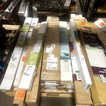 Stack of long, rectangular boxes with blinds or shades inside on a warehouse floor.